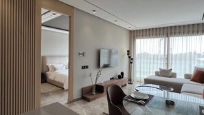A modern hotel room with a bed, a TV mounted on the wall, a dining area with a glass table, and a large window with curtains.