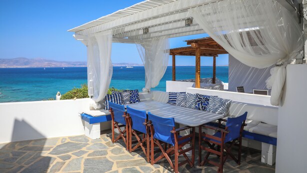 Une terrasse avec un sol en pierre, une table blanche et des chaises bleues, et une vue sur la mer.