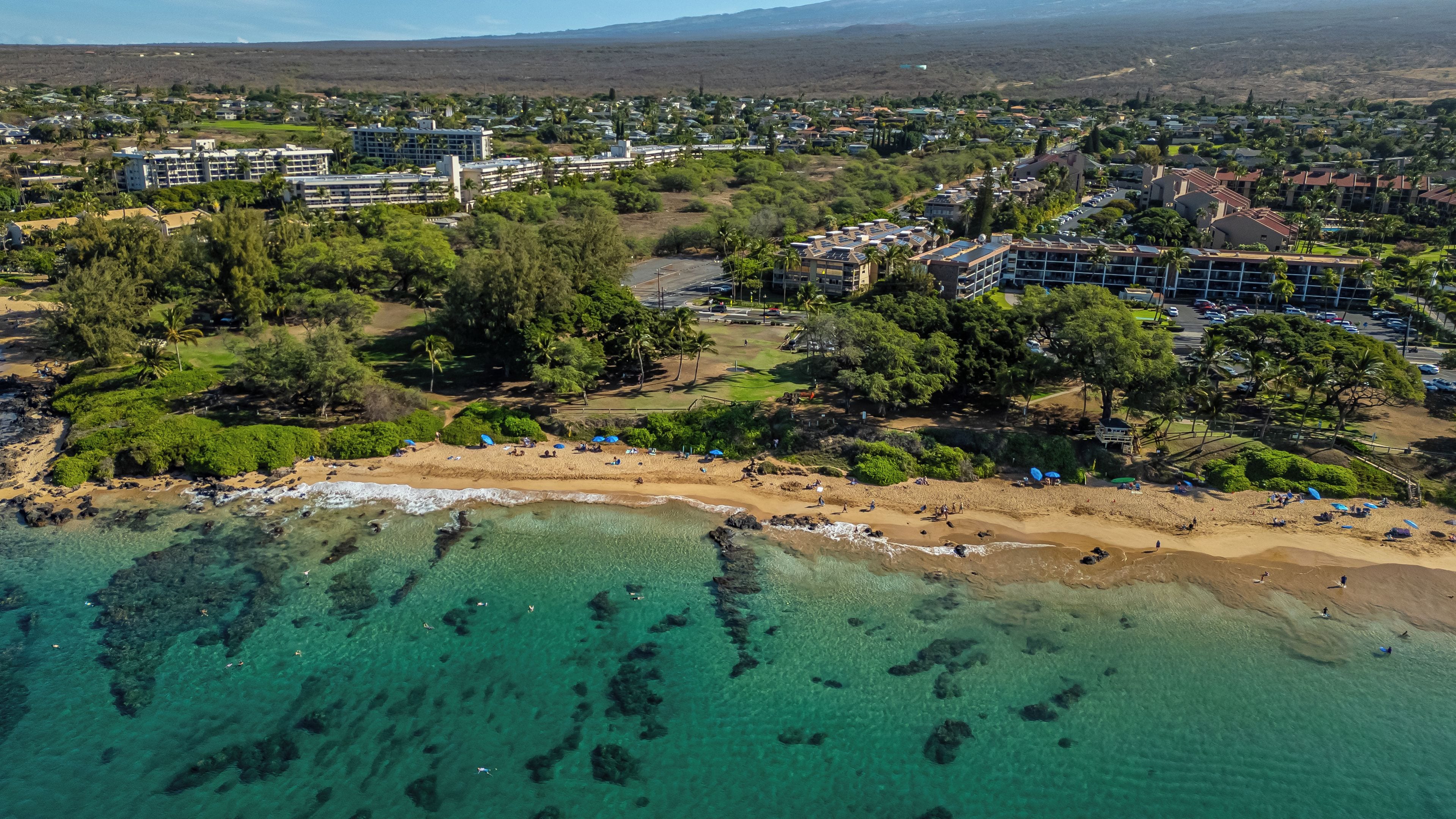 An aerial view of a coastal resort with multiple buildings, a sandy beach, and clear turquoise waters.