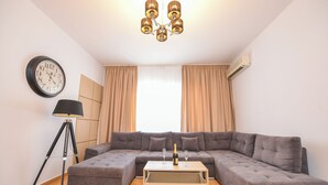 Uma sala de estar moderna com um sofá seccional, uma mesa de centro com uma garrafa e copos, um abajur de chão, um relógio de parede e um lustre.
