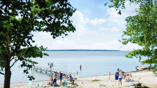 A beach with a pier extending into calm waters, surrounded by trees and people enjoying the area. - 4 Star Holiday Home in Farjestaden (Farjestaden)