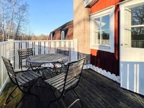 A wooden deck with a round table and chairs, a white fence, and a red building with a white door.