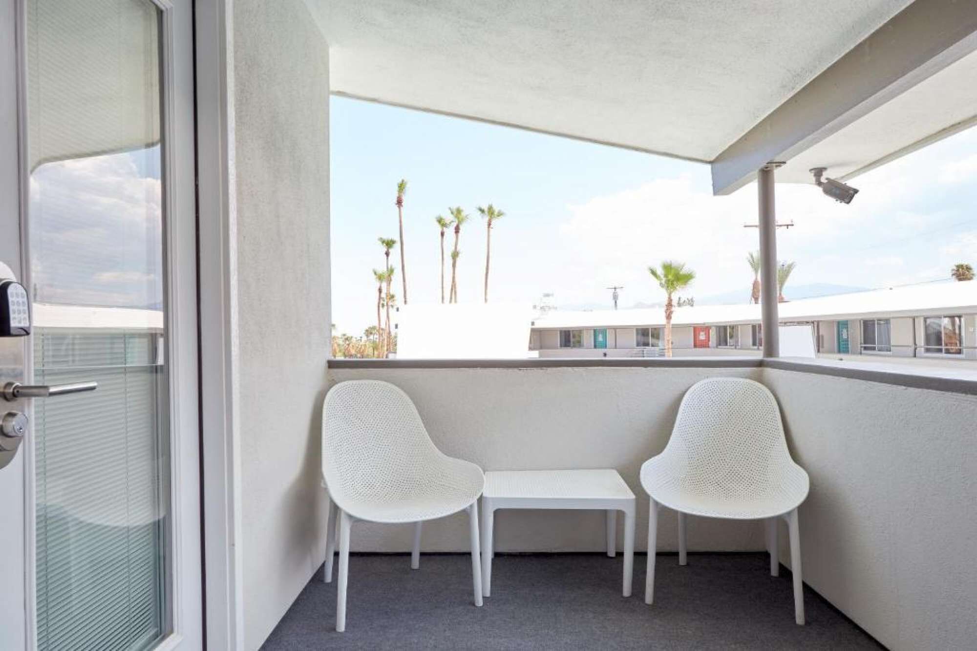 A balcony with two white chairs and a small table, overlooking a row of buildings and palm trees.