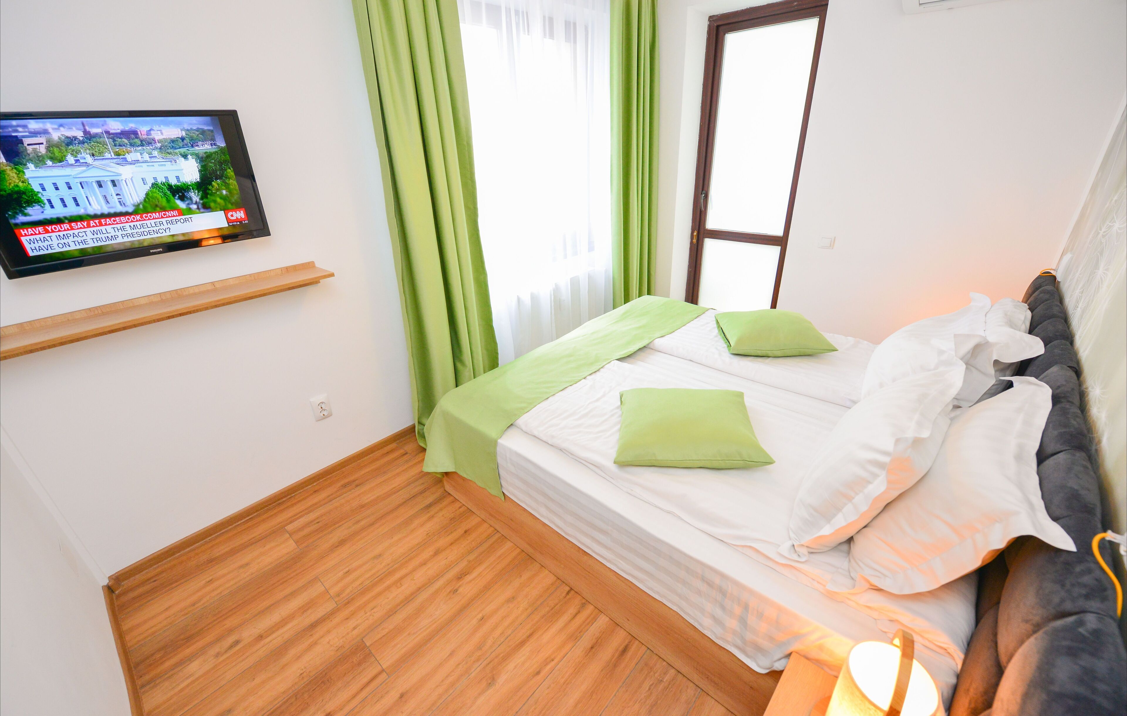 Una camera d'albergo con un letto, tende verdi, una televisione a parete e una finestra.
