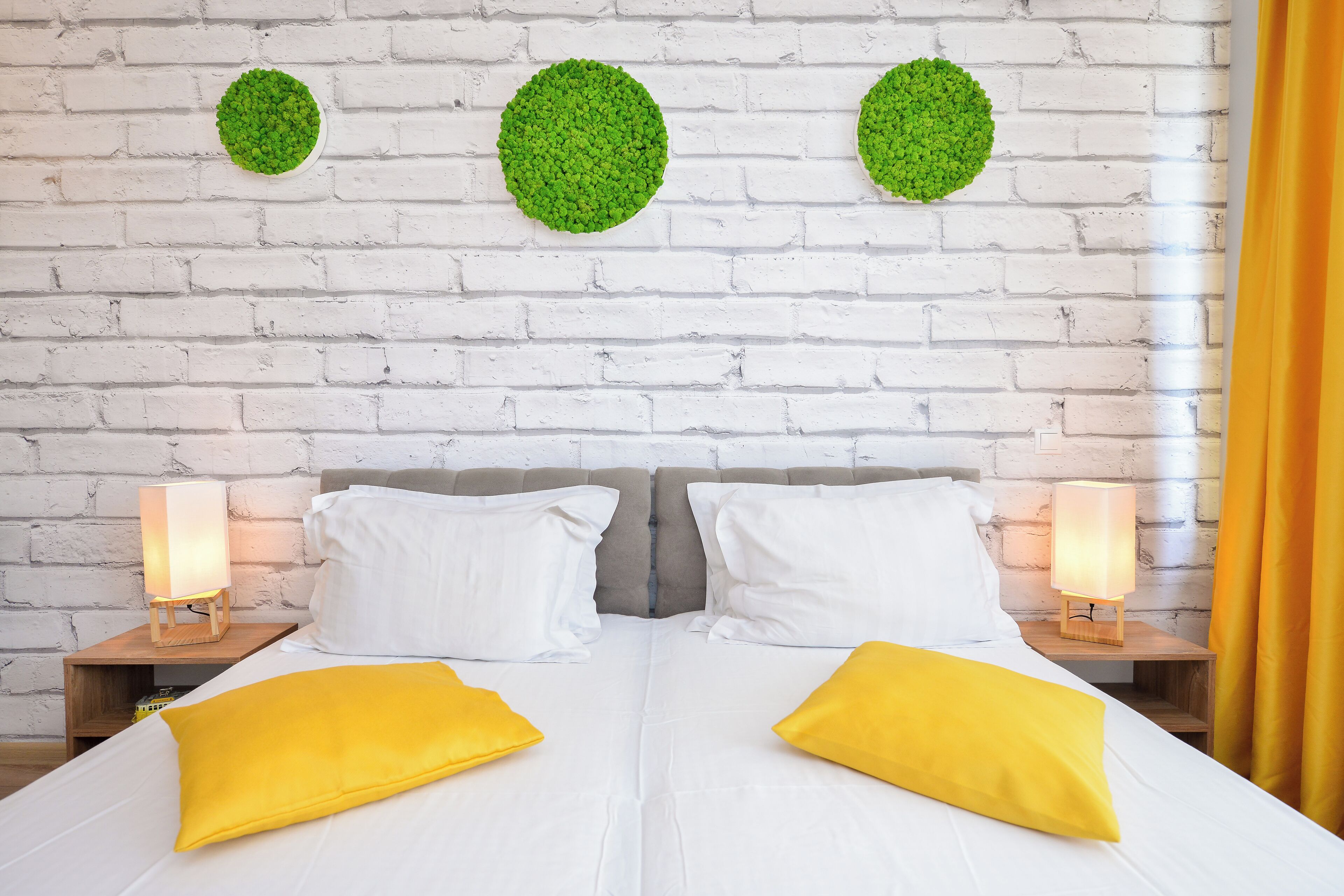 A bed with white bedding and two yellow pillows, flanked by two bedside tables with lamps, against a white brick wall with three green circular decorations.