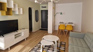A modern living room with a flat-screen TV, a sofa, a dining table with yellow chairs, and a patterned rug.