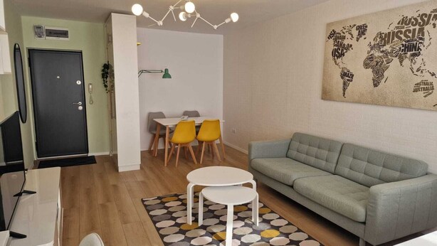 A modern living room with a grey sofa, a dining table with yellow chairs, a white coffee table, and a patterned rug.