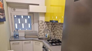 A compact kitchen with white cabinets, a yellow accent wall, a stainless steel refrigerator, and a window with a geometric pattern.