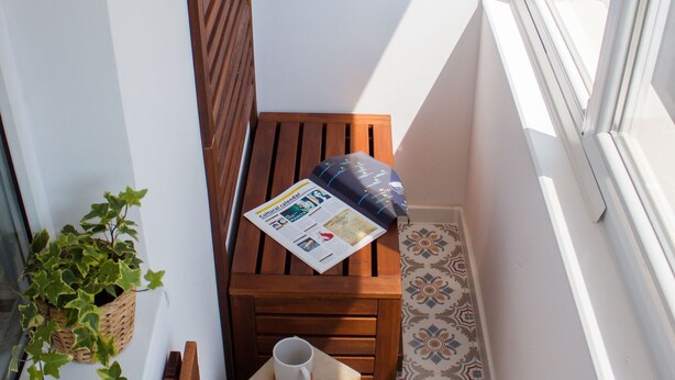 Un petit balcon étroit, avec un banc en bois, une petite table sur laquelle reposent un journal, une tasse et un pot de fleurs.