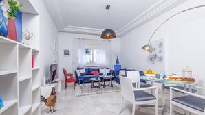 Un salon moderne avec un canapé bleu, des fauteuils blancs, une table à manger avec des chaises, et un coin cuisine à l’arrière-plan.