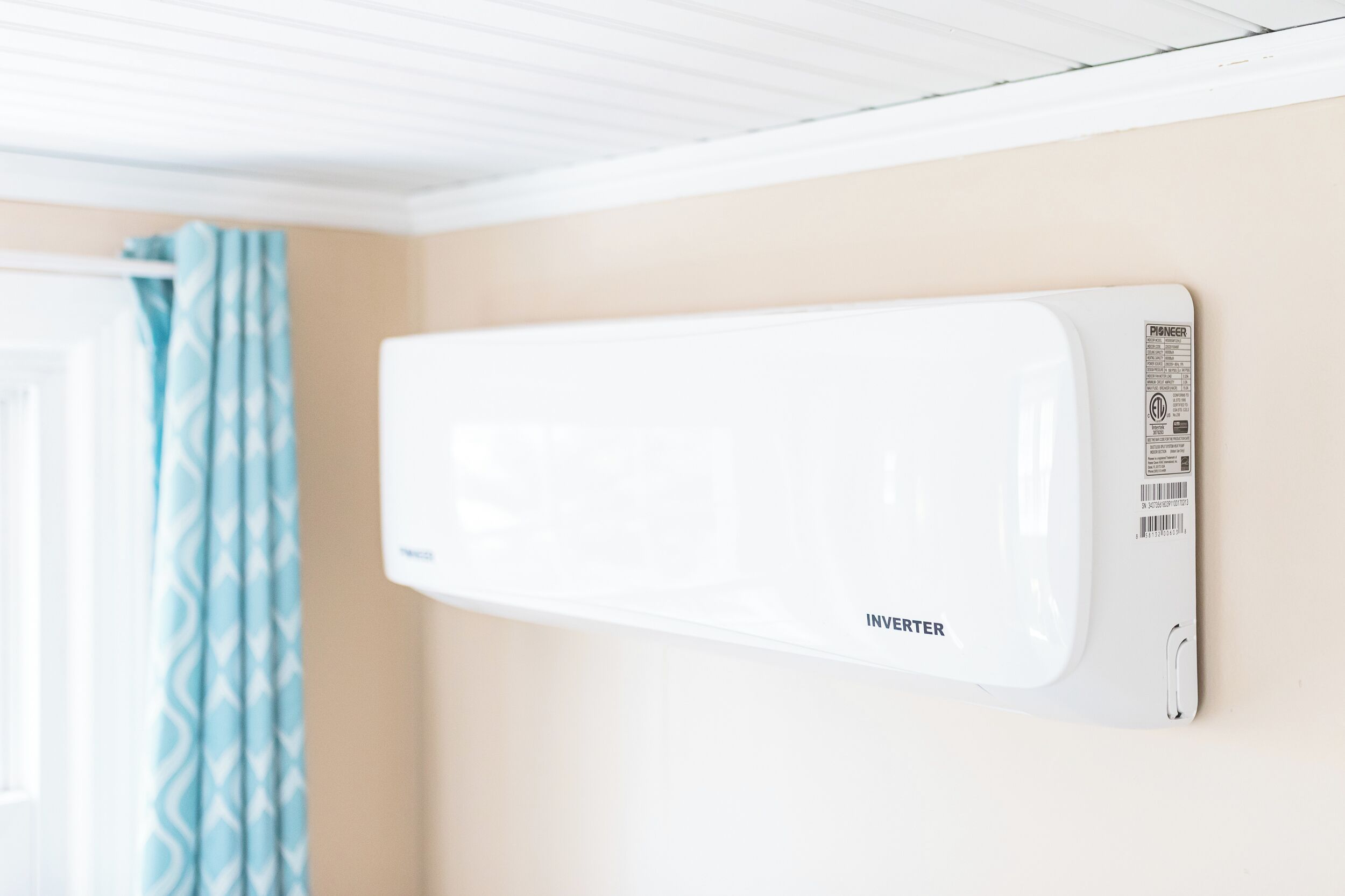 A white wall-mounted air conditioner with a Pioneer brand label, a window with a blue curtain, and a white ceiling with a wooden panel design.