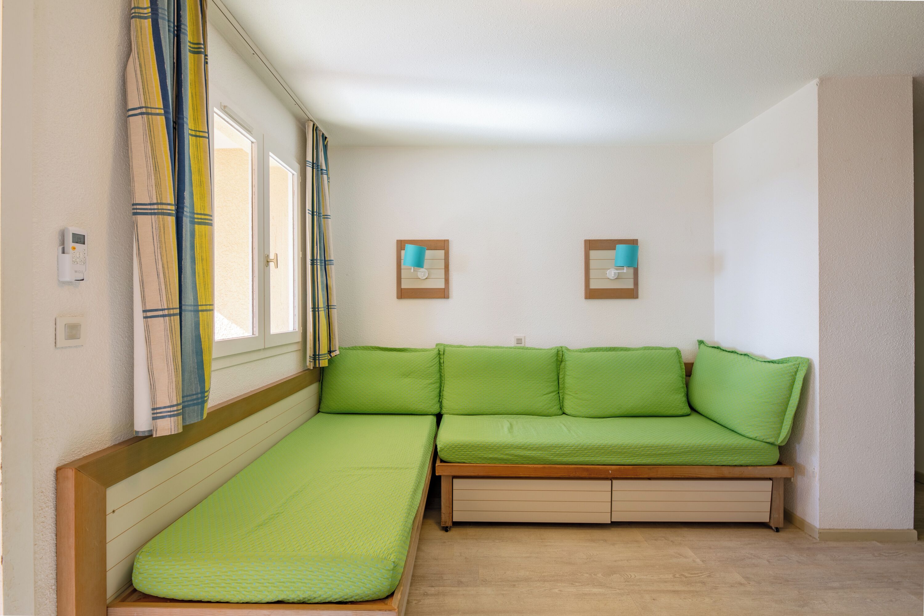 A compact room with a green sofa, a small bed, and two wall-mounted light fixtures.