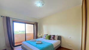 A bedroom with a bed, a nightstand, a window with curtains, and a view of greenery outside. - Albufeira Sunny Style by Homing (Albufeira)