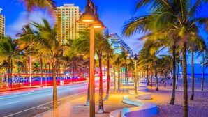 Un paseo marítimo frente a la playa con palmeras, farolas y una carretera con estelas de luz.