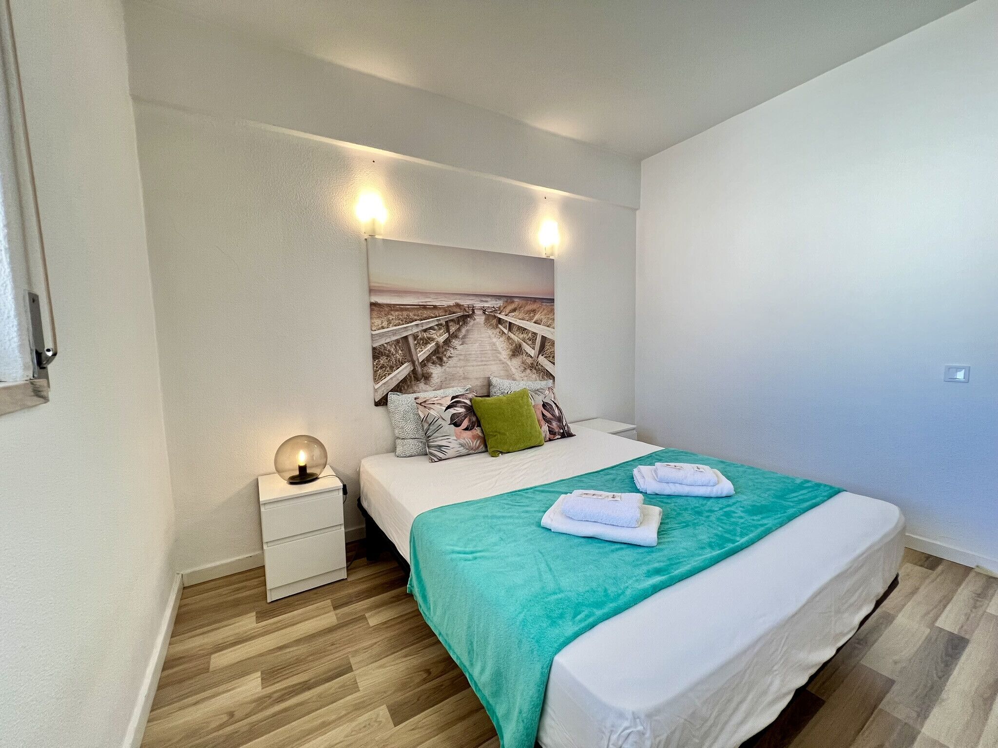 Un dormitorio con cama, mesita de noche, lámpara de pared y una obra de arte con motivos playeros.