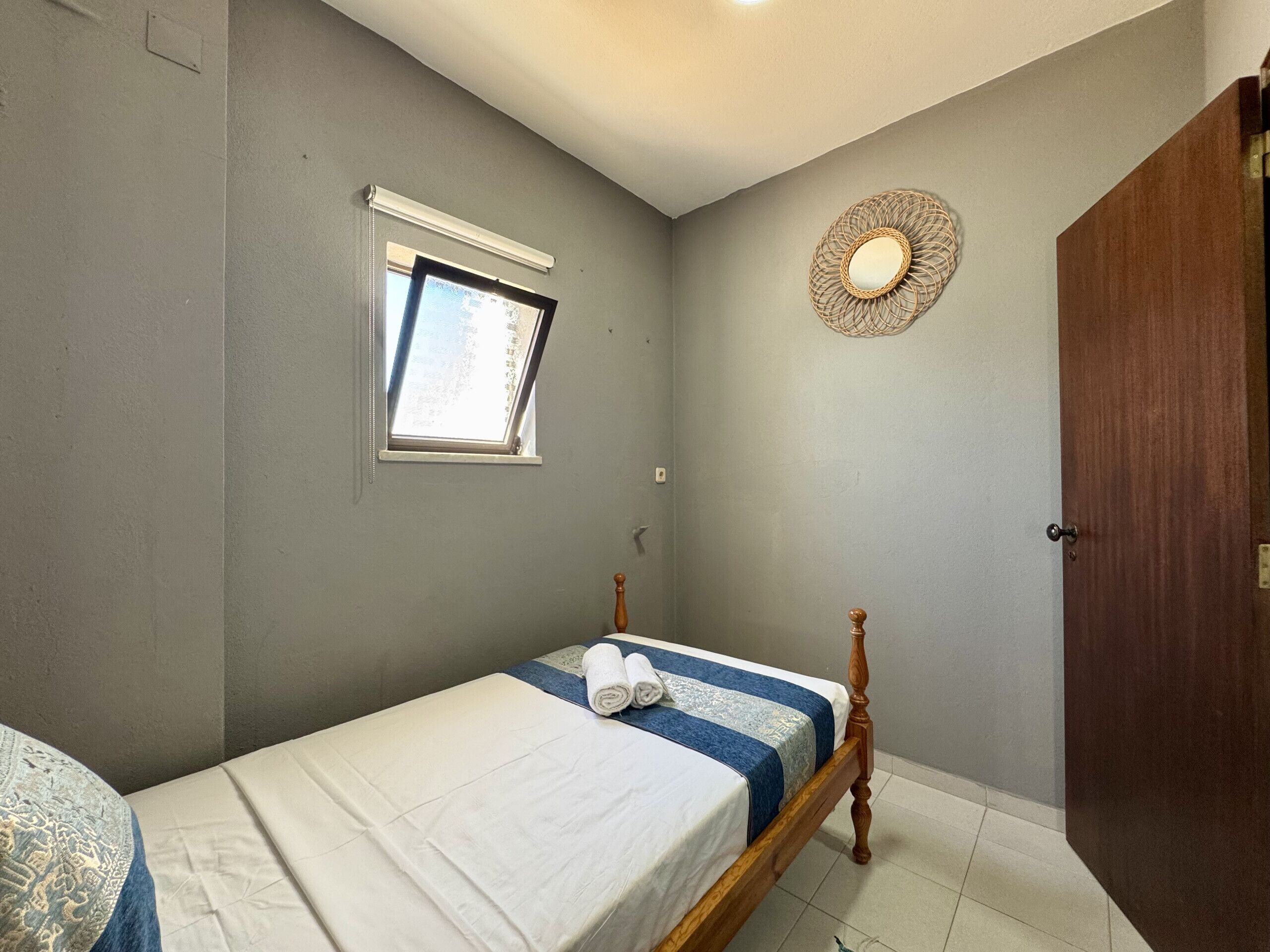 A small bedroom with a single bed, a window, a door, and a wall-mounted light fixture.