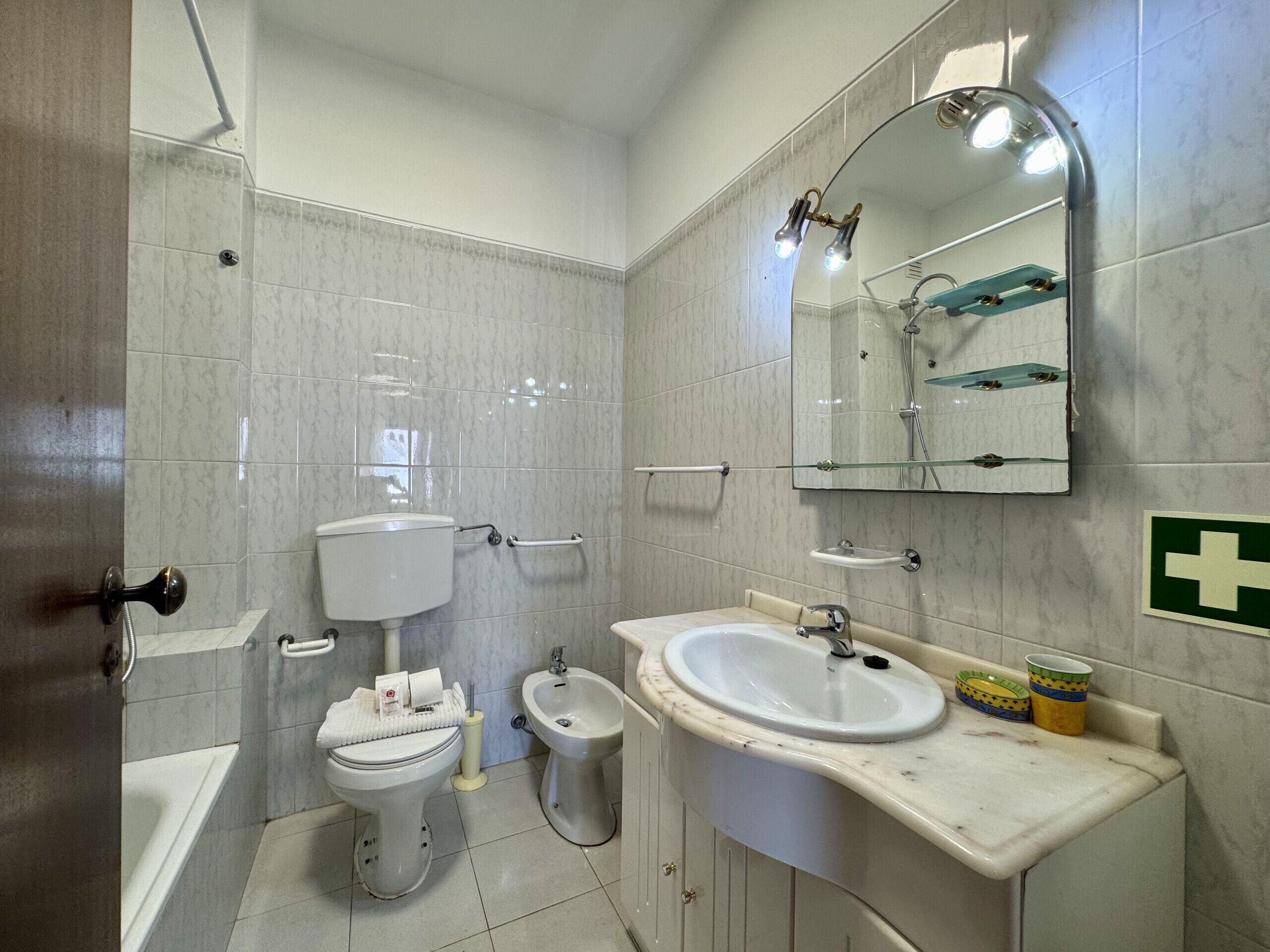 Un baño con inodoro, bidé, lavamanos y un espejo con repisa de vidrio.