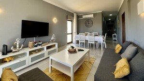 A living room with a TV, white furniture, a dining area with a clock, and a gray sofa. - Albufeira Central 8 by Homing (Albufeira)