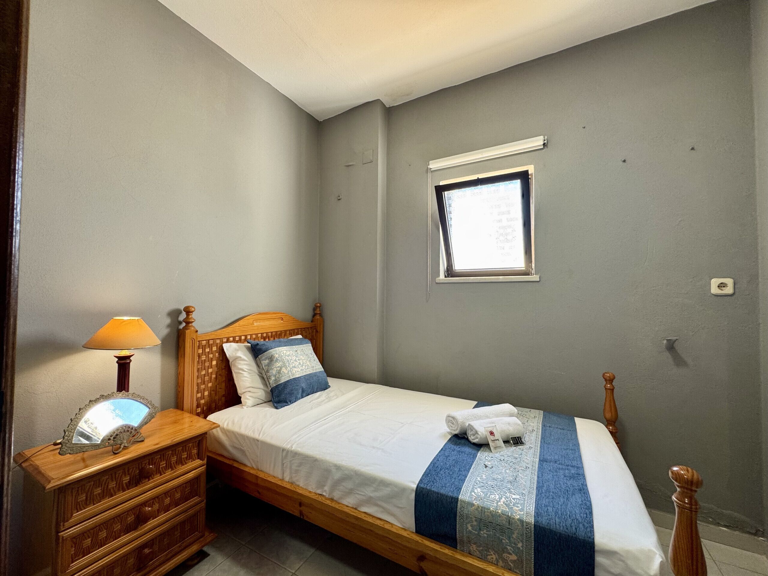 Un dormitorio con cama, mesita de noche, lámpara, ventana y una lámpara fijada a la pared.