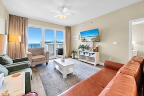 A spacious living room with a balcony, a flat-screen TV, and a view of the beach. - Ariel Dunes 1208 - Beautiful 2-bdrm w/ Gulf & pool views, steps to the beach! (Miramar Beach)