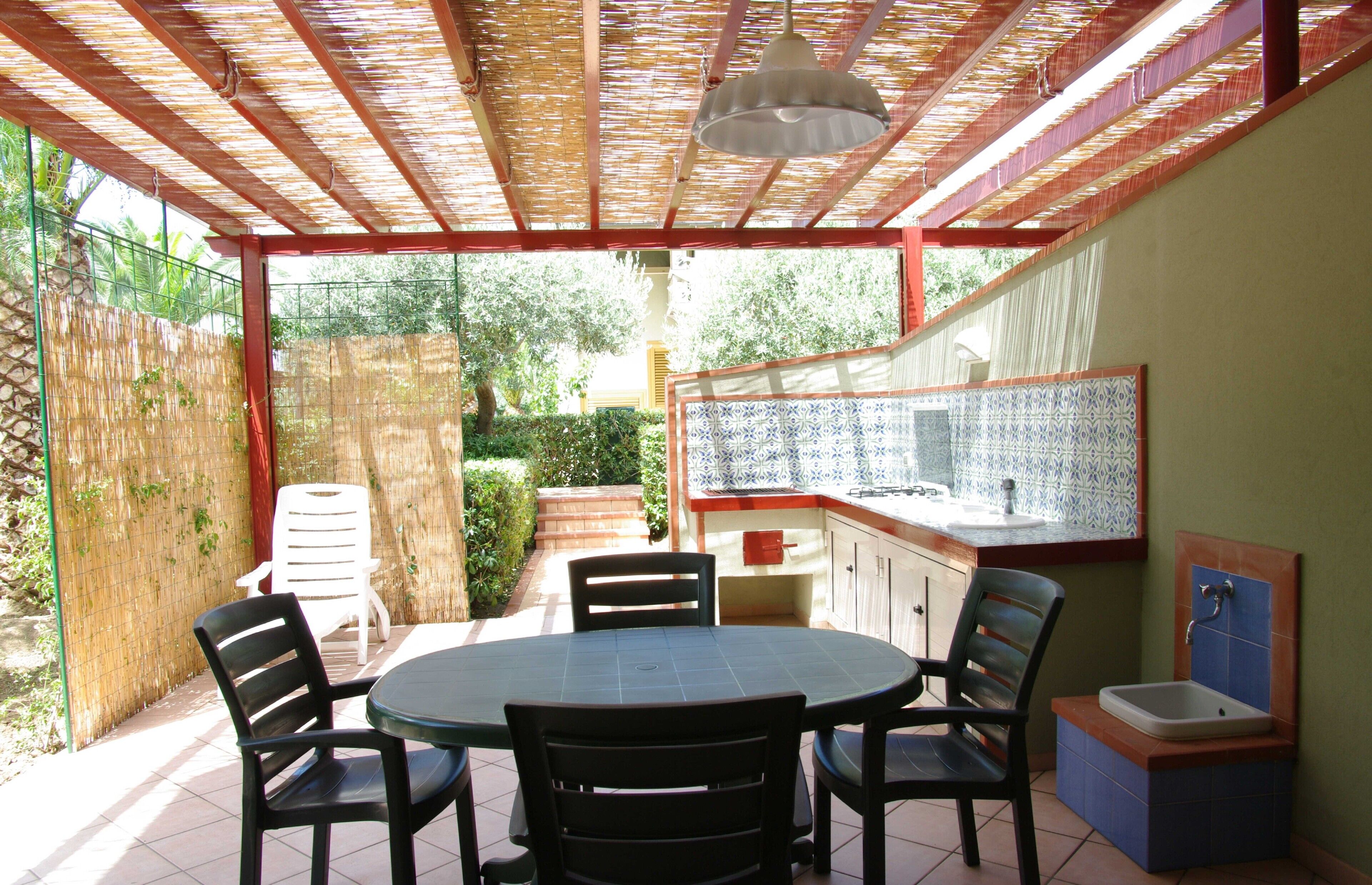 A covered patio with a round table and four chairs, a sink, and a tiled backsplash.