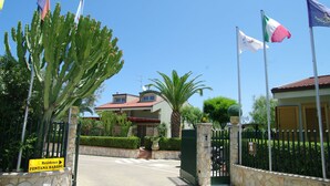 Una entrada protegida por una reja con un letrero que dice "Residence Fontana Barone" y varias banderas.