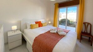 Una habitación de hotel con una cama grande, una mesita de noche con lámpara, una silla y vistas al exterior a través de una puerta corredera.