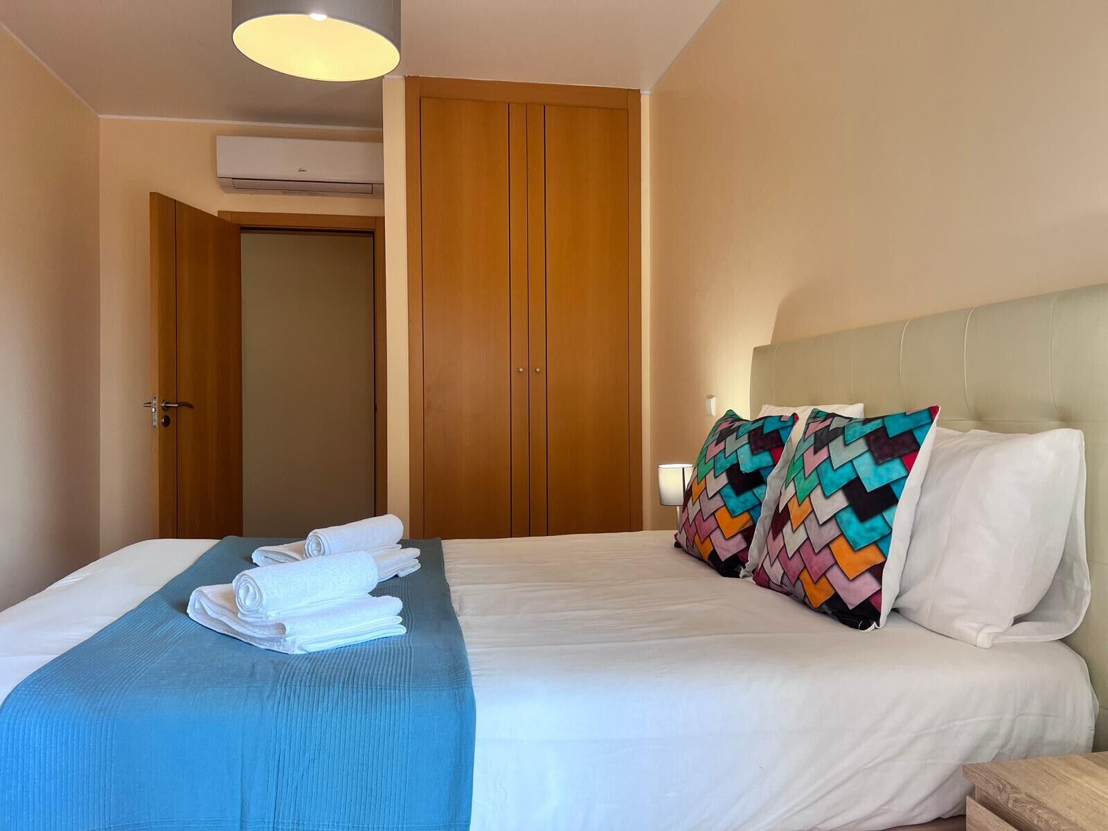 Quarto de hotel com cama, colcha azul, travesseiro branco e travesseiros coloridos.