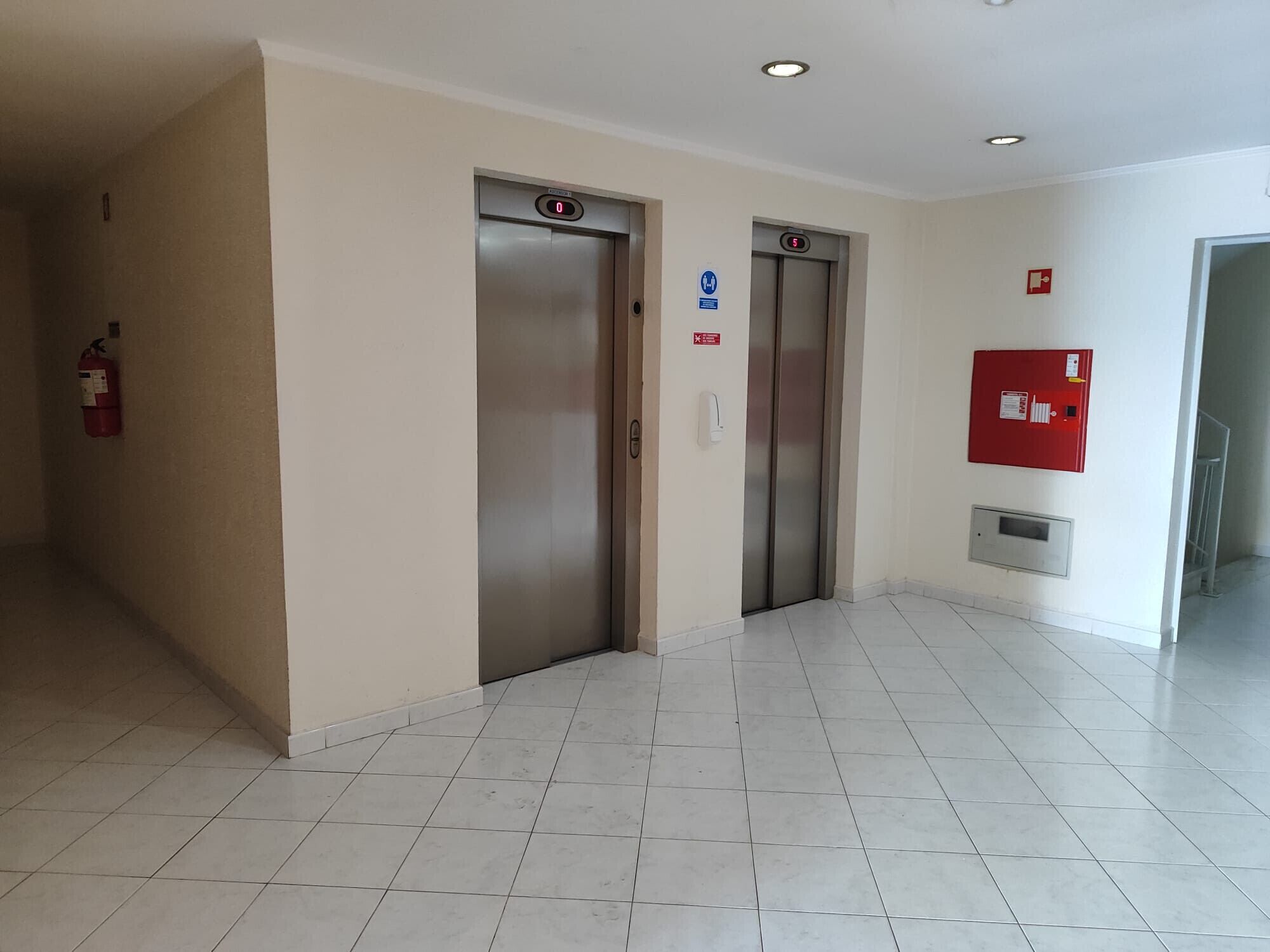 Un espace dédié aux ascenseurs, comprenant deux ascenseurs, un extincteur et un détecteur d’incendie fixé au mur.