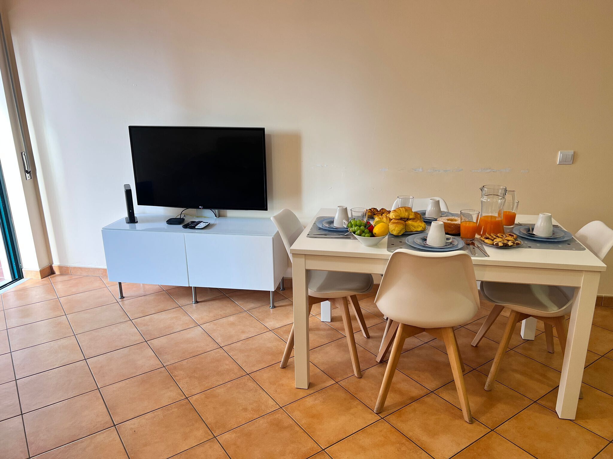 A dining area with a TV on a white cabinet, a table set with plates, glasses, and a bowl of fruit.