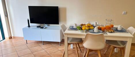 A dining area with a TV on a white cabinet, a table set with plates, glasses, and a bowl of fruit.