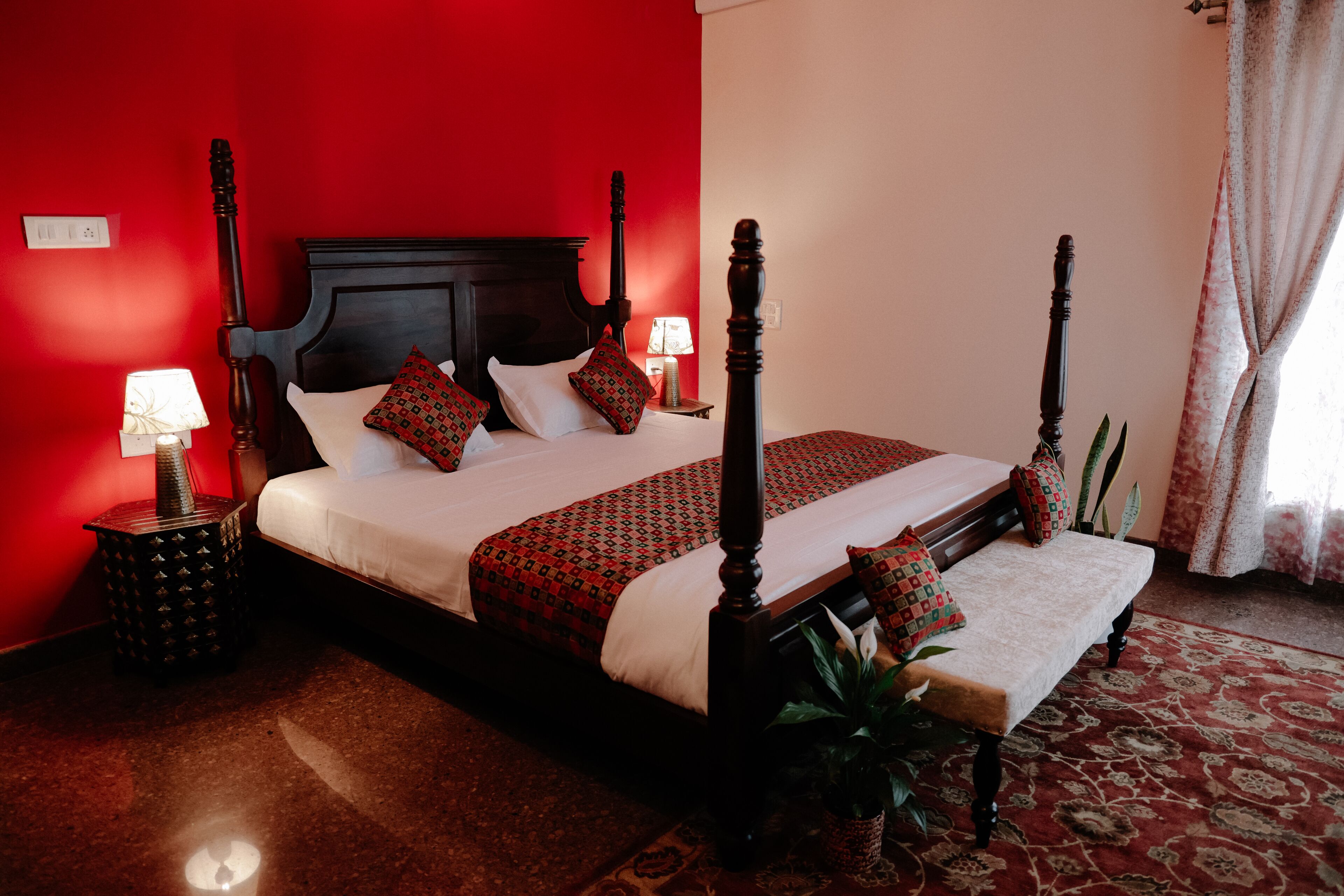 Een hemelbed met een donker houten hoofdbord, witte lakens en beddengoed met een rood en groen patroon. Een klein nachtkastje met een lamp en decoratief object is zichtbaar.