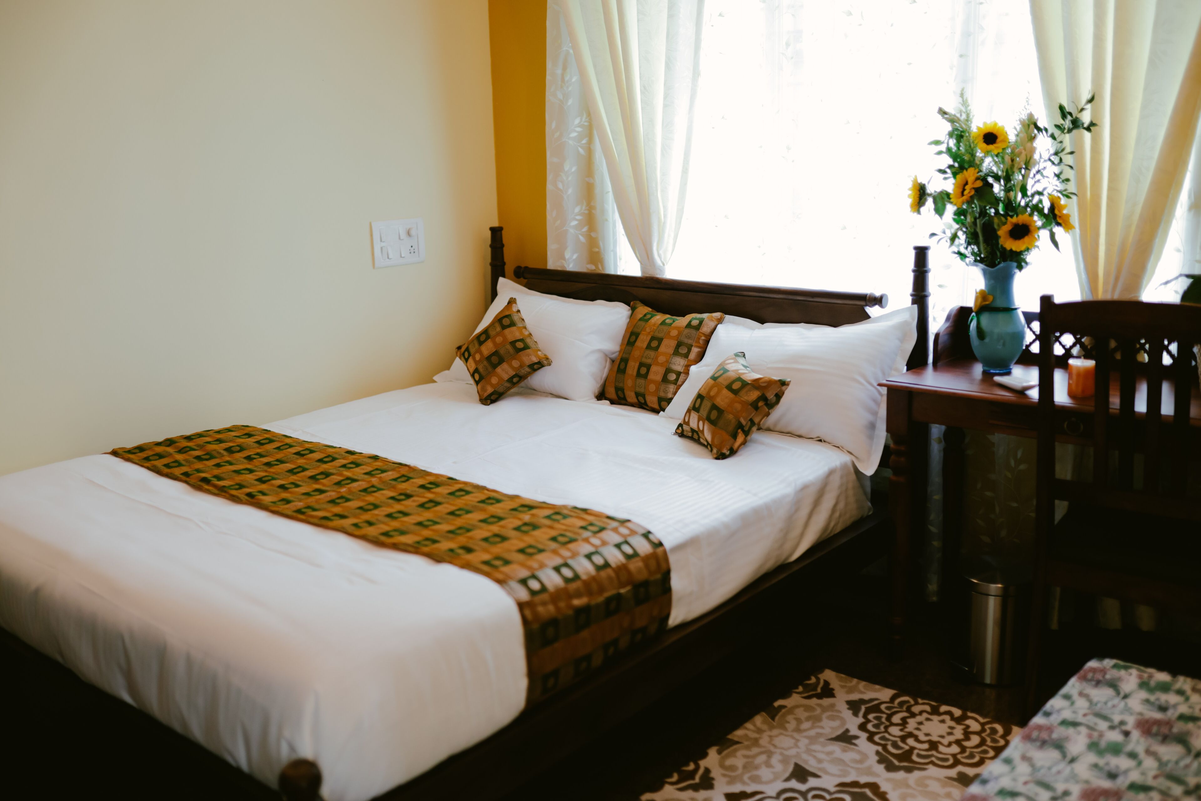 A neatly made bed with white linens and patterned pillows, a wooden headboard, and a vase of sunflowers on a nightstand.