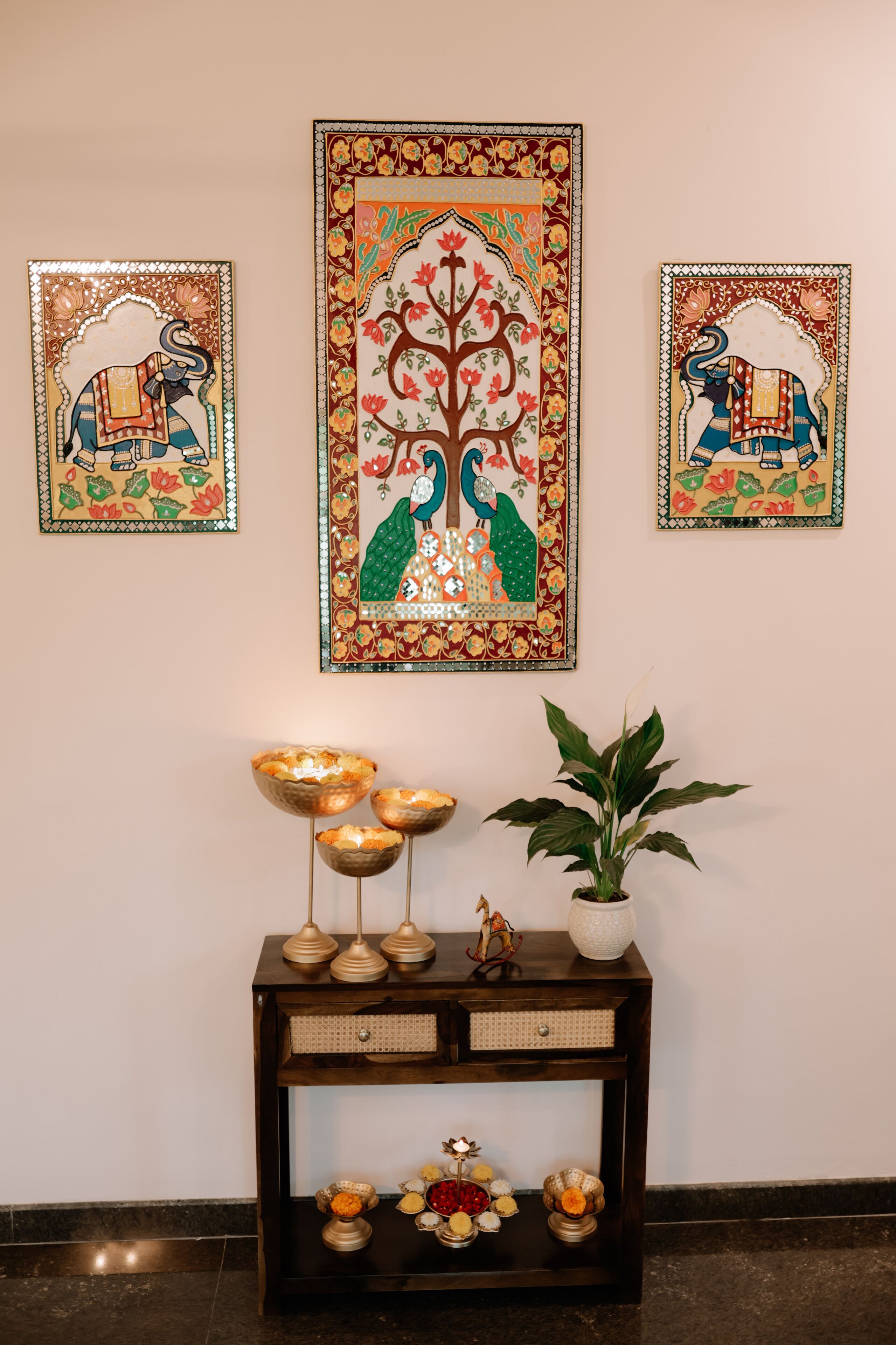 Een muur met drie ingelijste kunstwerken waarop olifanten en pauwen staan afgebeeld, een donker houten consoleschrank met decoratieve kommen en een potplant.