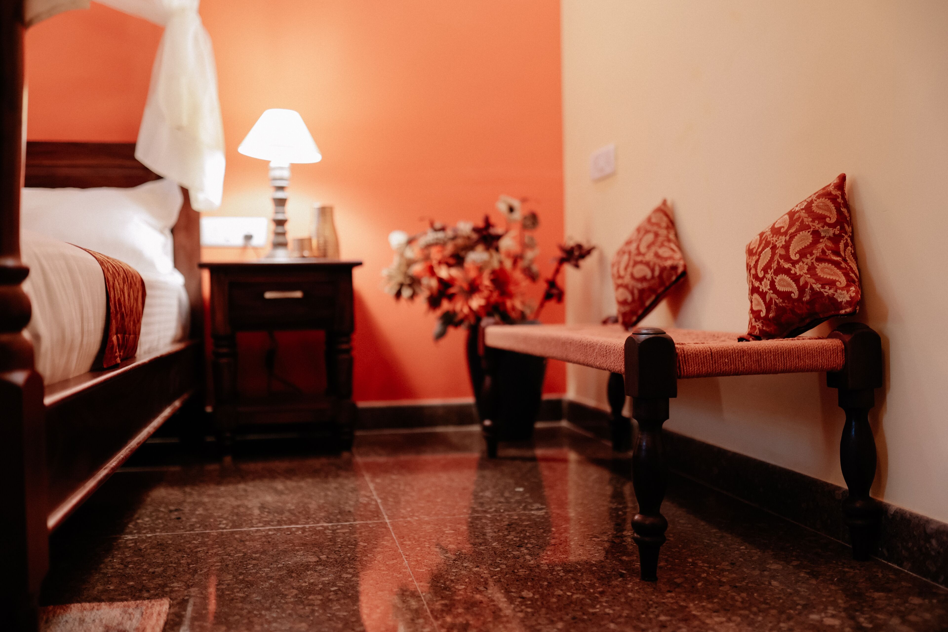 Quarto de hotel com cama, mesa de cabeceira com abajur, um banquinho com almofadas e um vaso com flores.