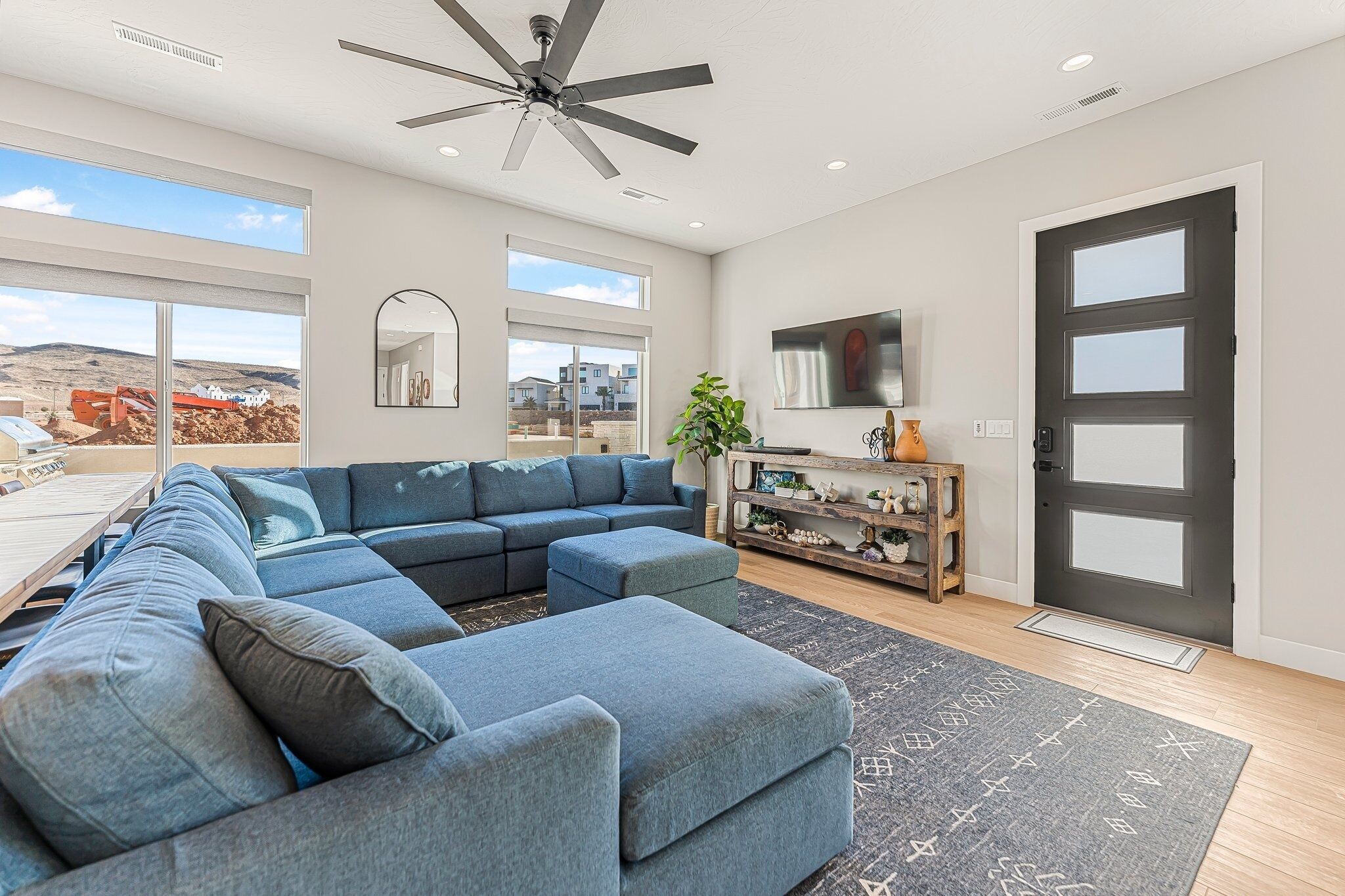 A modern living room with a sectional sofa, a ceiling fan, large windows, and a wooden console table.
