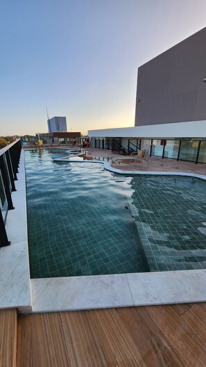 Una piscina amb zona de solàrium enrajolatada, envoltada d'edificis moderns i un cel serè.