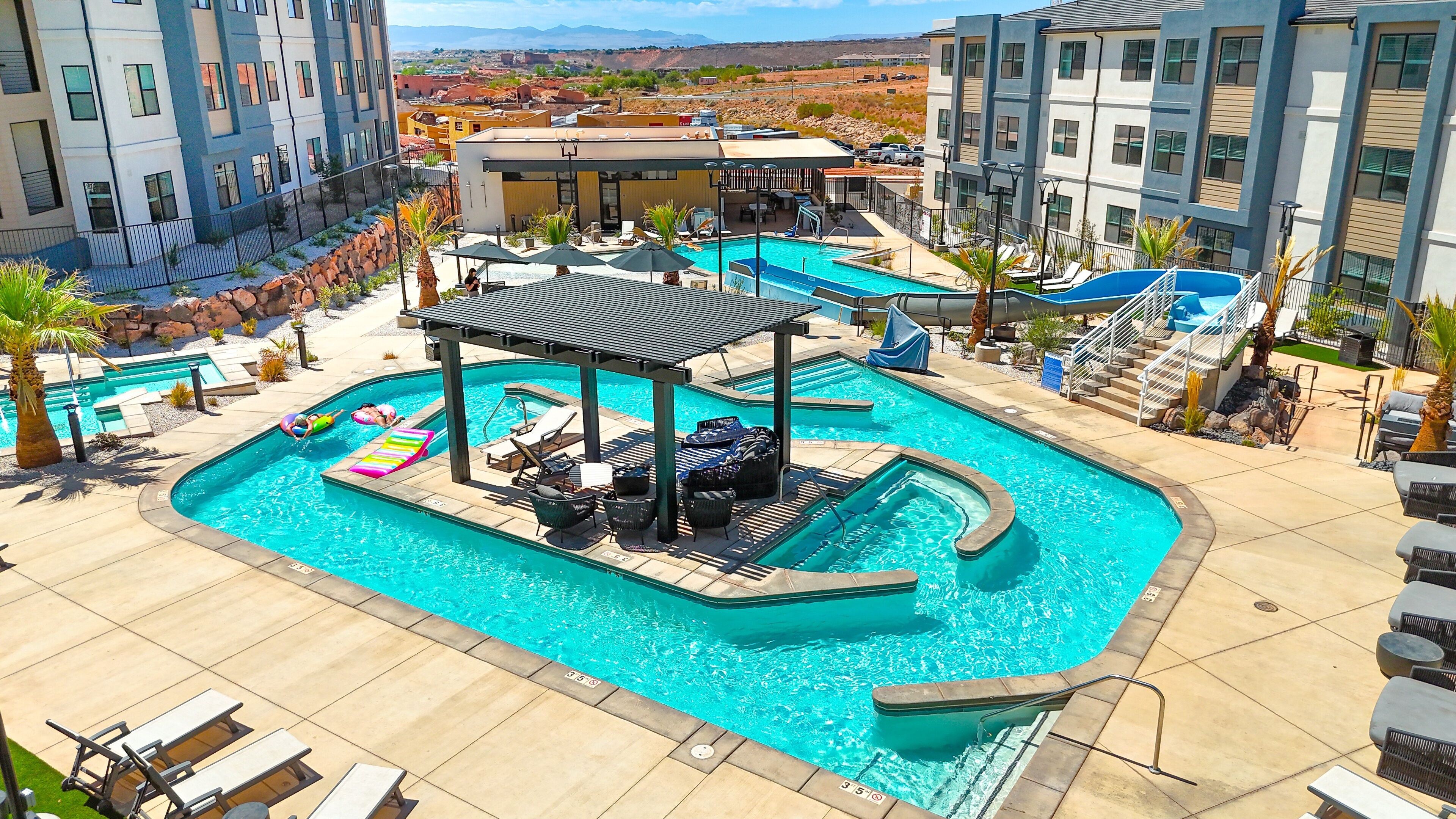 Una gran piscina al aire libre con tumbonas y una zona de estar cubierta, rodeada de edificios modernos y vistas al paisaje.