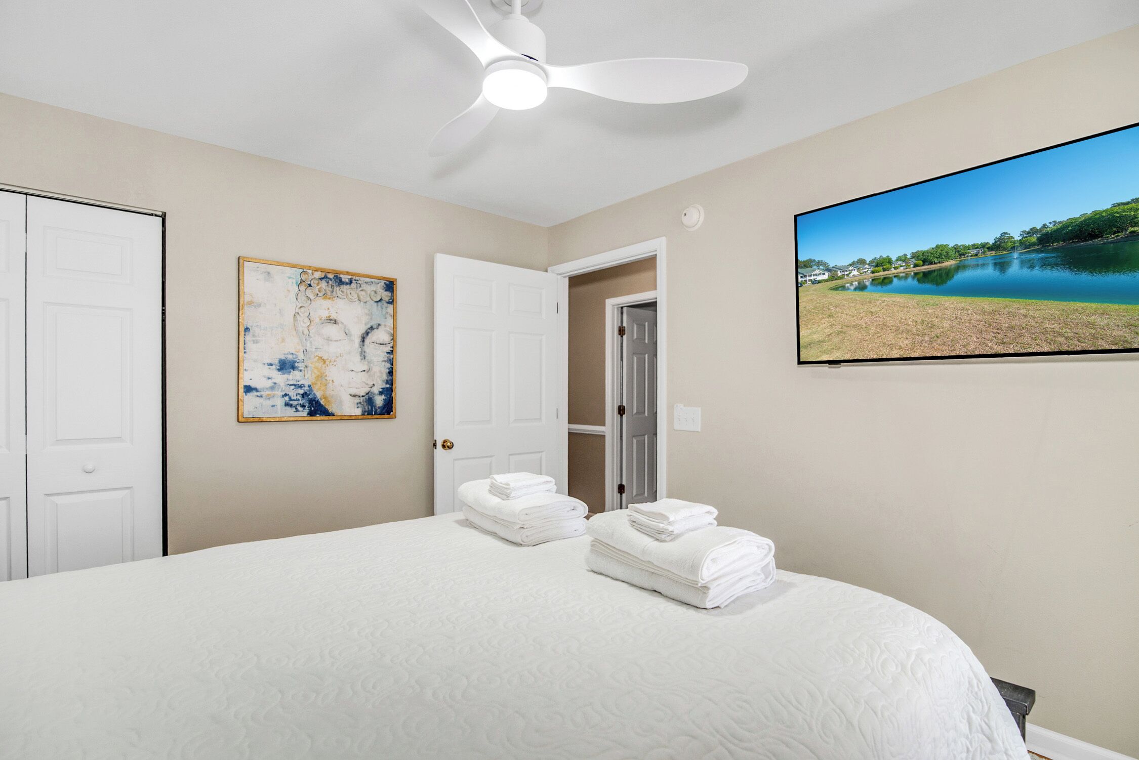 A bedroom with a bed, a ceiling fan, a painting, and a TV displaying a landscape.