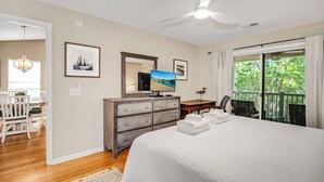 Un dormitorio con cama, escritorio, cómoda, televisor y balcón con vistas a árboles.