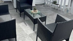 Un espace extérieur aménagé avec des meubles en osier et une table en verre, entouré de murs blancs et d’une clôture blanche en bois.