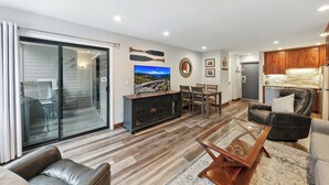 A modern living room with a glass sliding door, a flat-screen TV, a wooden cabinet, leather sofas, and a kitchen area in the background.