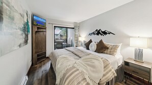 A bedroom with a bed, a TV mounted on the wall, a wooden cabinet, and a nightstand with a lamp.