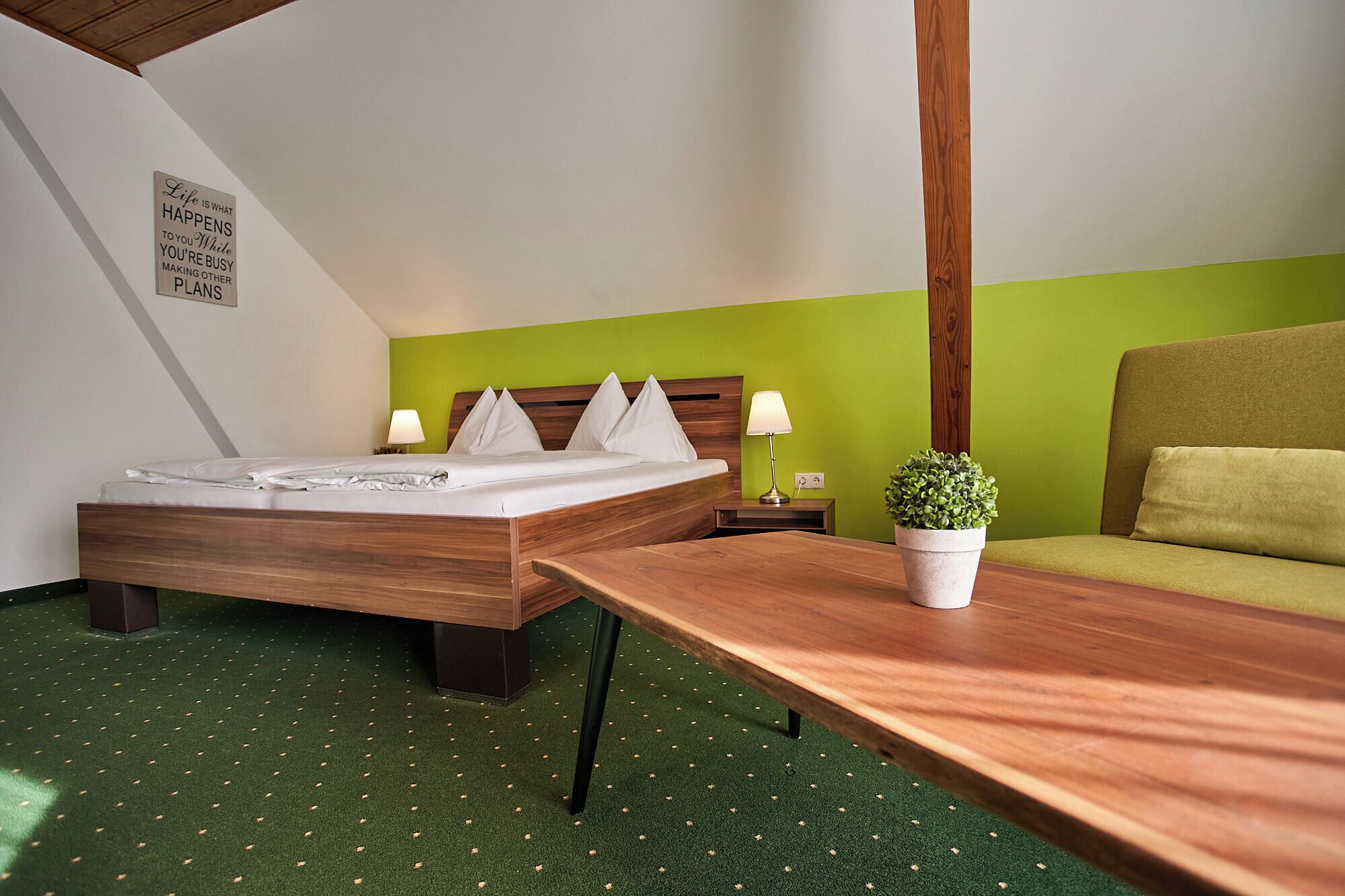 Una habitación de hotel con dos camas, una pared verde, una mesa de madera, un sofá verde y una planta en maceta.