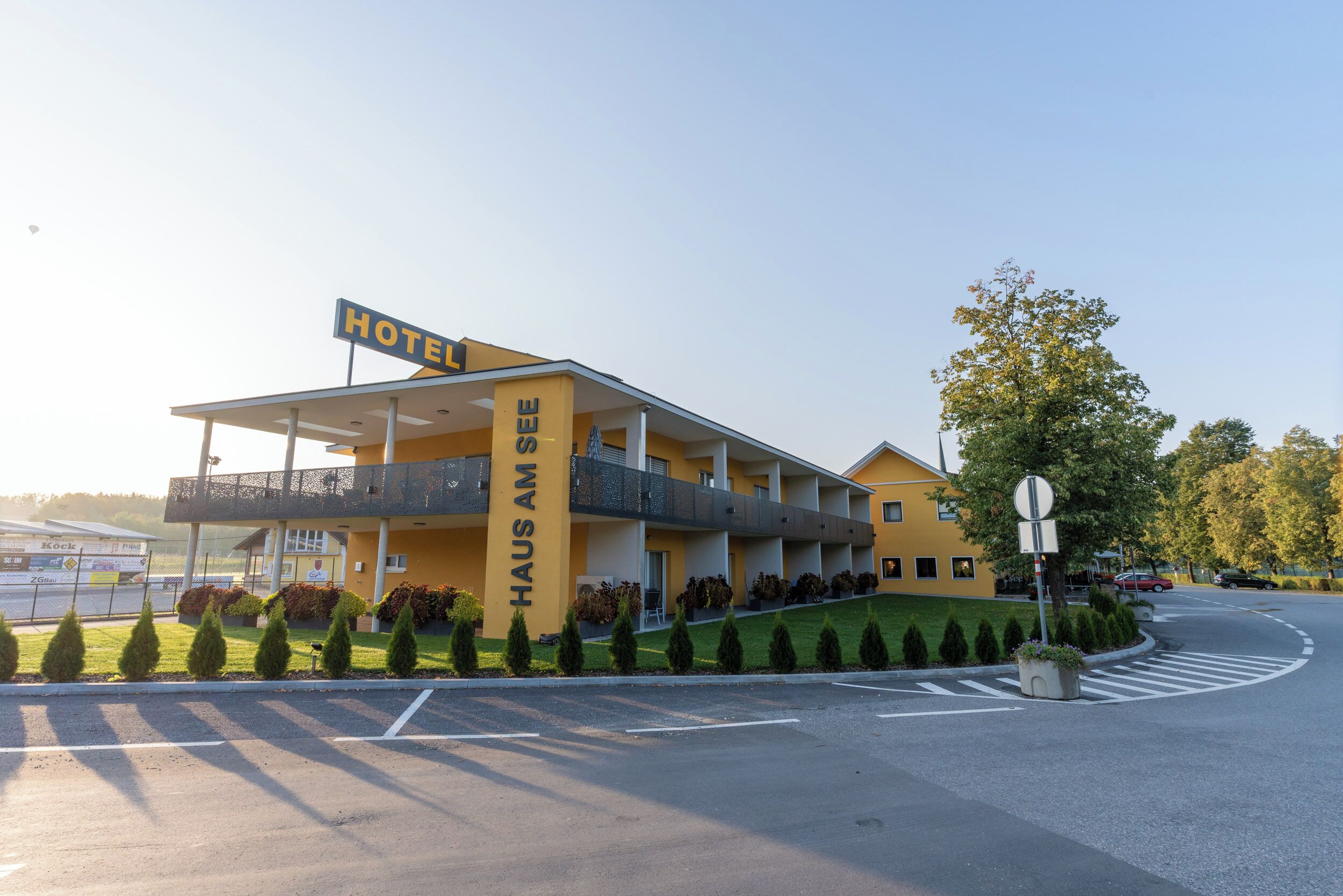 A hotel with a yellow facade, a sign reading "HAUS AM SEE," and a parking area with marked lanes.