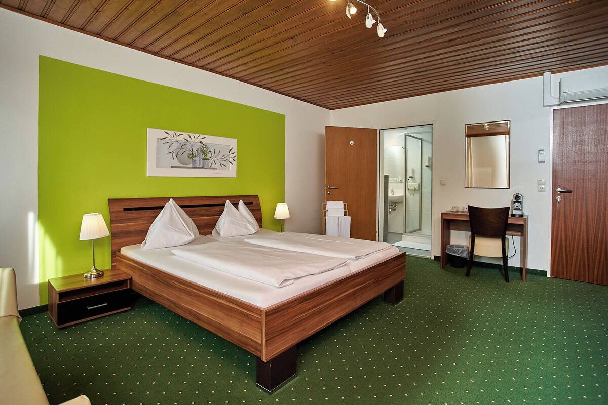 Ein Hotelzimmer mit einem hölzernen Bett, einem Schreibtisch, einem Stuhl, einem Spiegel und einem durch eine Glastür einsehbarer Badezimmer.