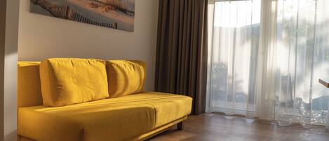 Un sofá amarillo en una habitación con suelo de madera, un cuadro en la pared y una ventana con cortinas.