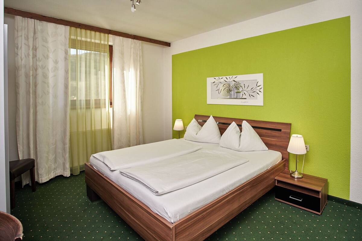 Ein ordentlich bezogenes Bett in einem Zimmer mit grüner Wand, einem Fenster mit transparenten Vorhängen und einem kleinen Nachttisch mit einer Lampe.