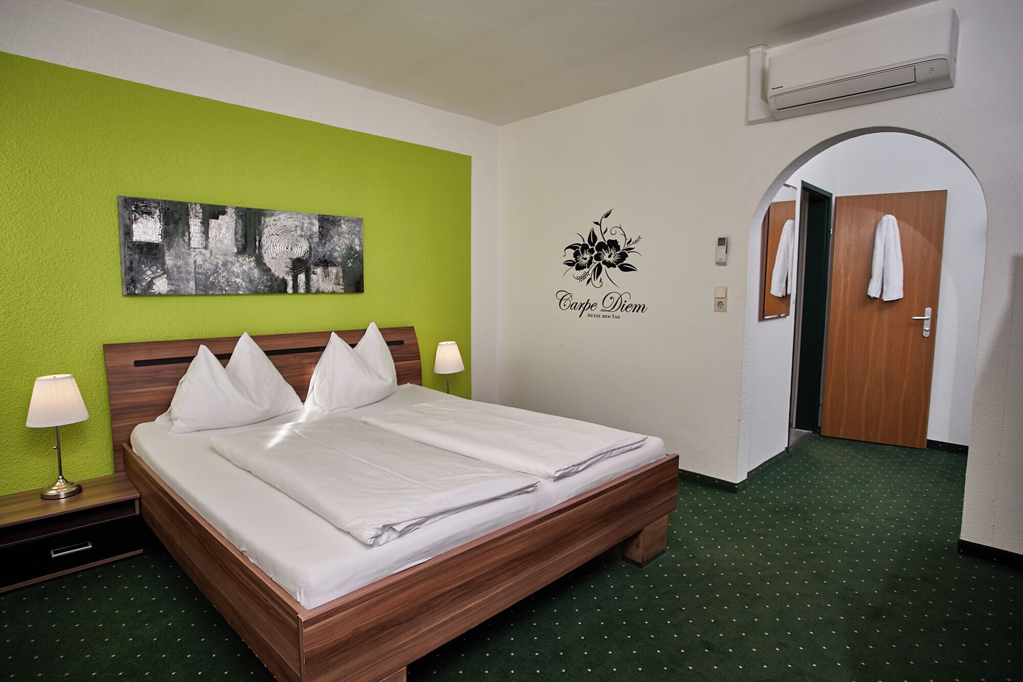 Ein Hotelzimmer mit Bett, grünen Wänden, einer Schwarz-Weiß-Grafik, einem Wandaufkleber mit der Aufschrift 'Carpe Diem', einem Handtuchhalter und einer Klimaanlage.