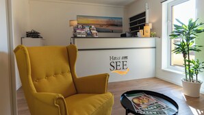 A yellow armchair, a reception desk with a beach painting, a potted plant, and a magazine on a tray. - Haus am See Stubenberg (Stubenberg)
